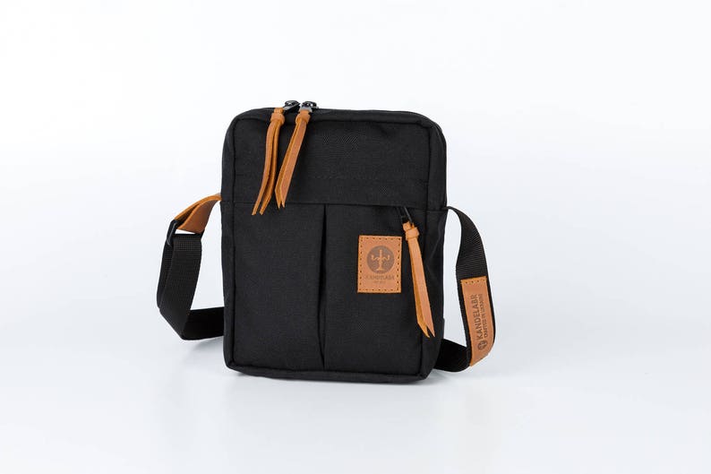 May include: A black canvas crossbody bag with brown leather accents and a zip closure. The bag has a small pocket on the front with a brown leather patch that says "LANDMADE" in black lettering.