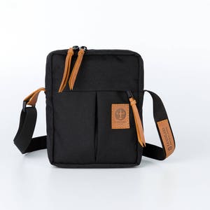 May include: A black canvas crossbody bag with a brown leather strap and zipper pulls. The bag has a front pocket with a brown leather patch that says "The Friendly Swede".