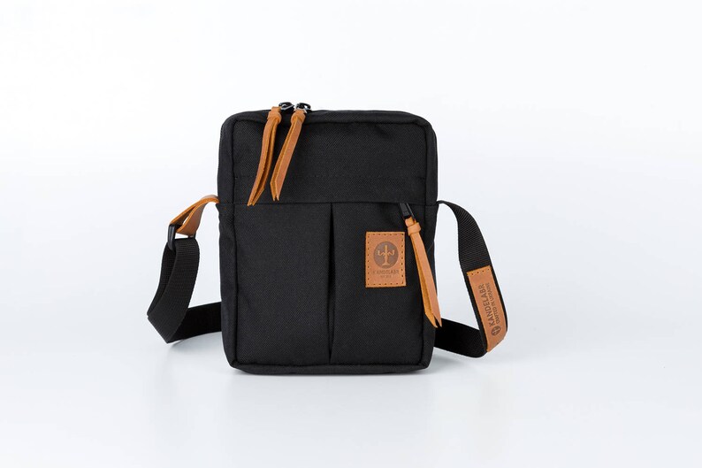 May include: A black canvas crossbody bag with a brown leather strap and zipper pulls. The bag has a front pocket with a brown leather patch that says "The Friendly Swede".