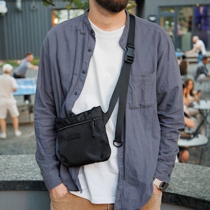 May include: A person wearing a blue button-down shirt over a white t-shirt and brown pants. They are wearing a black crossbody bag with a zippered pocket.