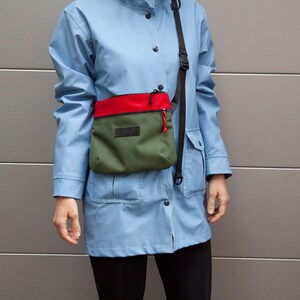 May include: A person wearing a light blue jacket with a green and red crossbody bag. The bag has a black zipper and a black strap.