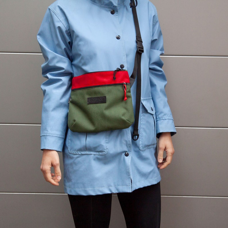 May include: A person wearing a light blue jacket with a green and red crossbody bag. The bag has a black zipper and a black strap.
