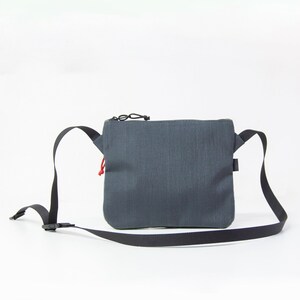 May include: A dark gray crossbody bag with a black adjustable strap and a zipper closure. The bag has a small red pull tab on the zipper.