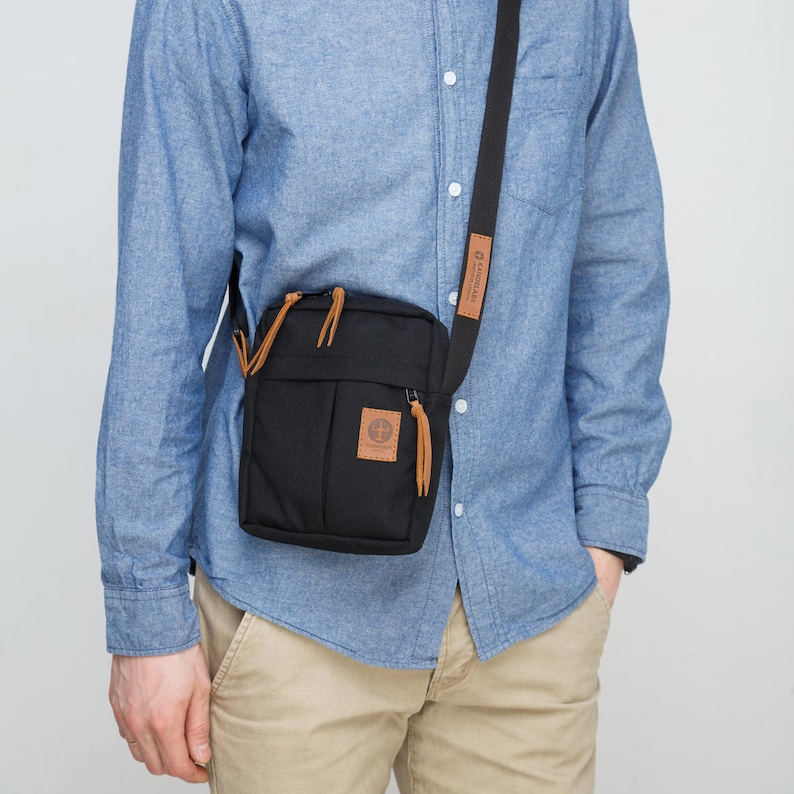 May include: A black canvas crossbody bag with a brown leather strap and zip closure. The bag has a small pocket on the front with a brown leather patch that says "Land's End".