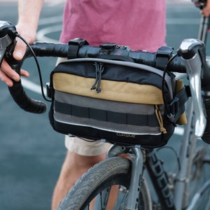 May include: A black handlebar bag with a tan and gray striped design attached to a bicycle handlebar. The bag has a zipper closure and the text "CURSUR" printed on the front.