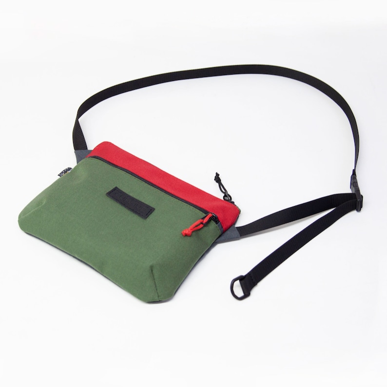 May include: A green and red canvas crossbody bag with a black adjustable strap. The bag has a zipper closure and a small pocket on the front.