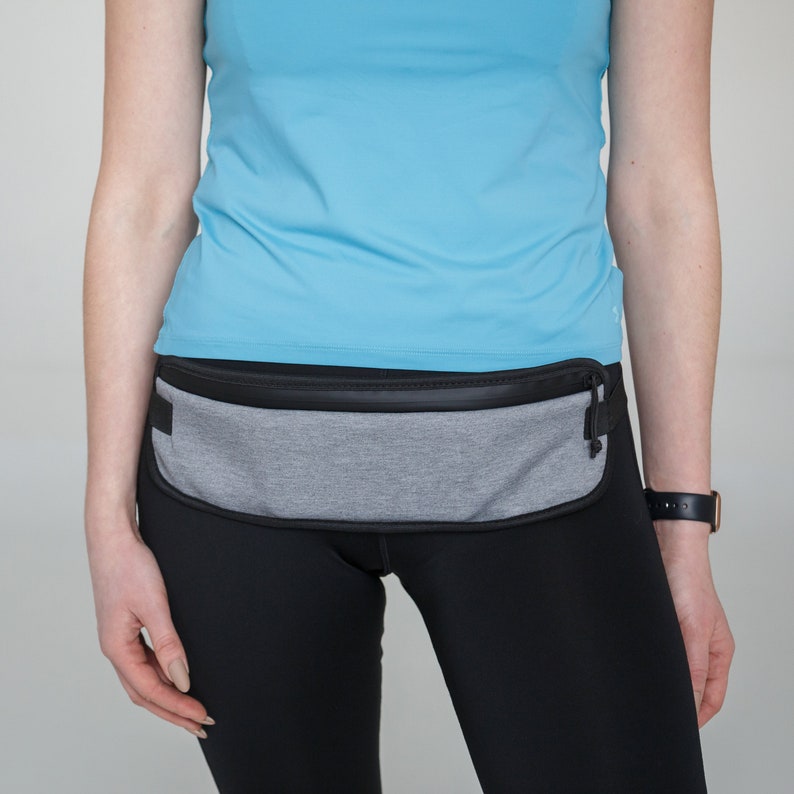 May include: A gray and black running belt with a zippered pocket. The belt is worn over a light blue shirt and black leggings.