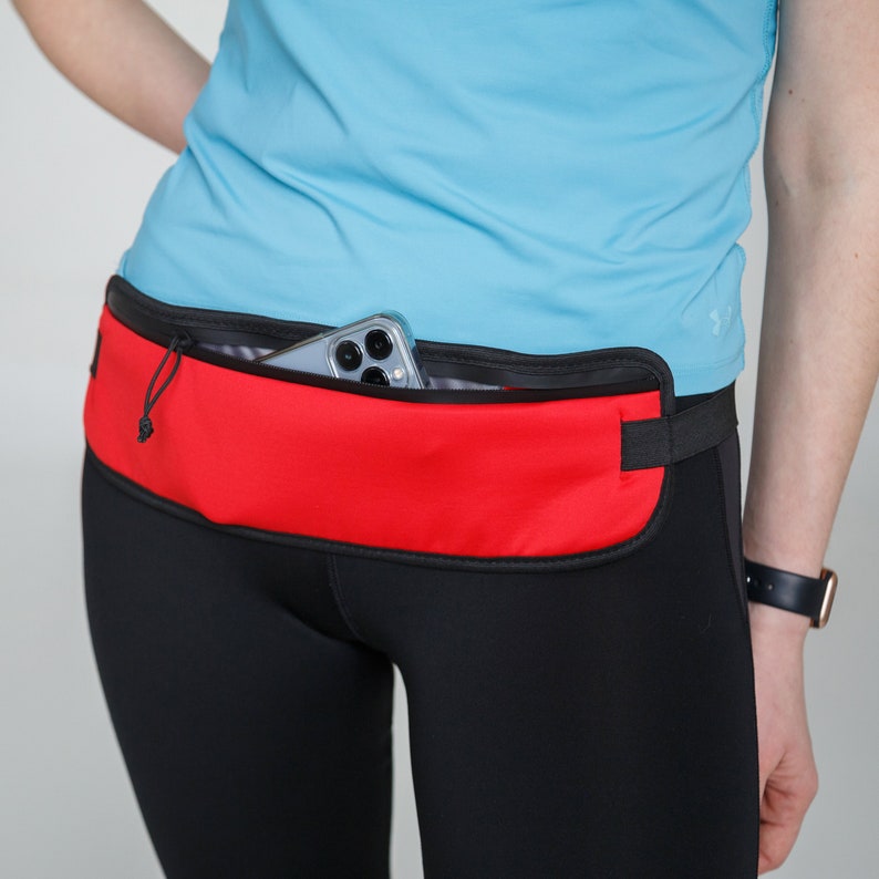 May include: A red running belt with a black zipper and a black adjustable strap. A smartphone is tucked into the pocket of the belt.