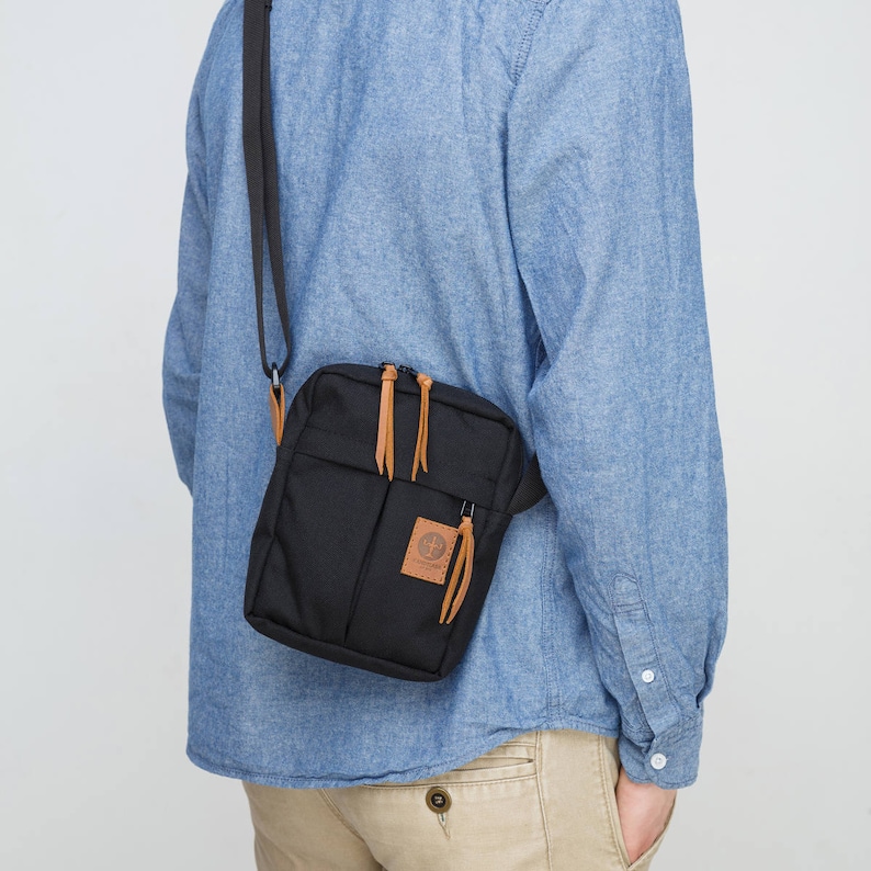 May include: A black crossbody bag with a brown leather strap and zipper closure. The bag has a small pocket on the front with a brown leather logo.