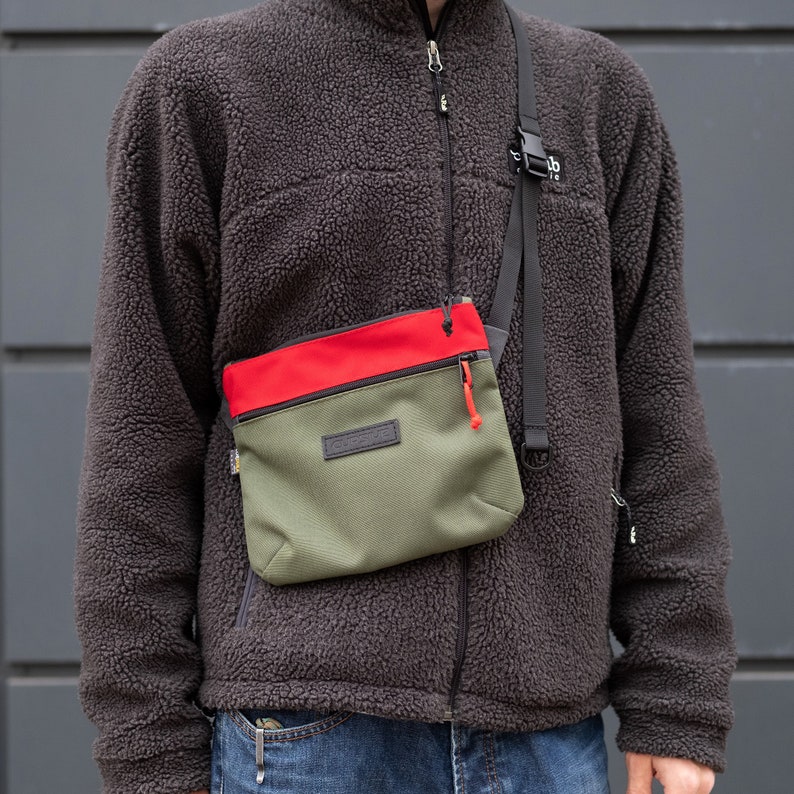 May include: A person wearing a brown fleece jacket with a red and green crossbody bag. The bag has a zipper closure and a small pocket on the front. The bag has the word "OUTDOOR" printed on the front.