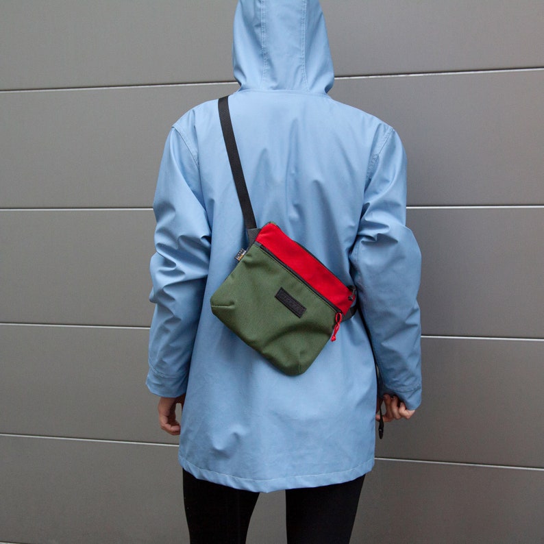 May include: A person wearing a blue hooded jacket with a green and red crossbody bag. The bag has a black strap and a small black label.
