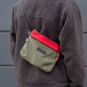 May include: A green and red crossbody bag with a black zipper and a black logo that says "CURT" on the front.