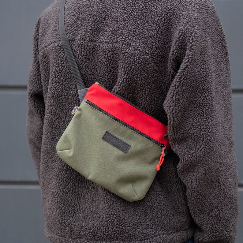 May include: A green and red crossbody bag with a black zipper and a black logo that says "CURT" on the front.