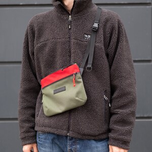 May include: A person wearing a brown fleece jacket with a zipper and a small green, red, and black crossbody bag with a zipper. The bag has a black strap and a silver buckle.