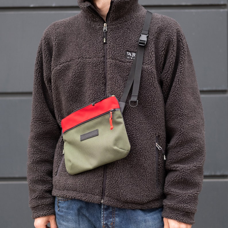 May include: A person wearing a brown fleece jacket with a zipper and a small green, red, and black crossbody bag with a zipper. The bag has a black strap and a silver buckle.