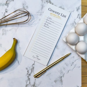 Peut inclure: Un bloc-notes de liste d'épicerie blanc avec un surlignage jaune et les mots "Grocery List" écrits en noir. La liste comprend des articles tels que des œufs, des bananes, du beurre, du lait et du pain. Un stylo doré et une banane sont également visibles sur l'image.