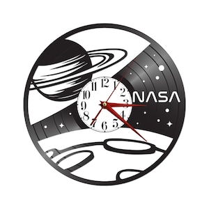 May include: Black and white wall clock with a space theme. The clock face is a vinyl record with a planet and moon design. The word "NASA" is printed on the right side. Red clock hands.