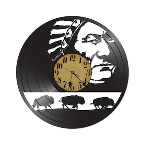 May include: Black vinyl record clock with a white silhouette of a Native American chief wearing a headdress. The clock face is in the center of the record and has a gold background with black numbers. Three white silhouettes of buffalo are at the bottom of the record.