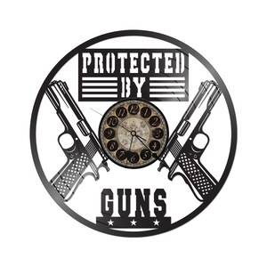 Puede incluir: Un reloj de pared circular negro con una esfera de estilo vintage. El reloj presenta dos siluetas de pistolas y las palabras "PROTECTED BY GUNS". Las manecillas del reloj señalan aproximadamente las 7:00.