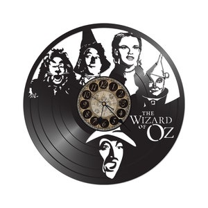 May include: Black vinyl record clock featuring characters from the Wizard of Oz, including Dorothy, the Scarecrow, the Tin Man, the Cowardly Lion, and the Wicked Witch of the West. The clock face is white with black numbers and hands. The text "The Wizard of Oz" is printed in white on the black vinyl.