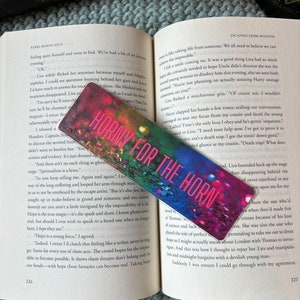 May include: A pink and red bookmark with the text "Horny for the Horn" on it. The bookmark is made of paper and has a floral design. The bookmark is lying on top of an open book with the title "Escaping from Houdini" on the cover.