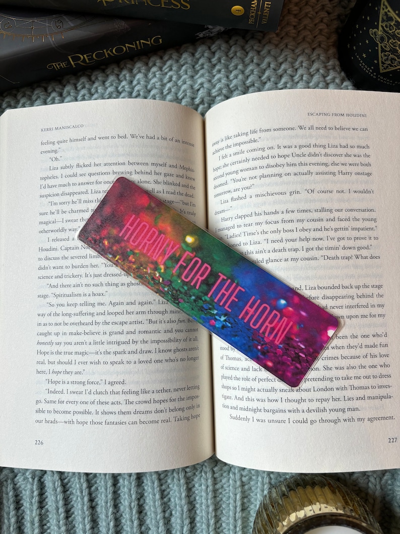 May include: A pink and red bookmark with the text "Horny for the Horn" on it. The bookmark is made of paper and has a floral design. The bookmark is lying on top of an open book with the title "Escaping from Houdini" on the cover.
