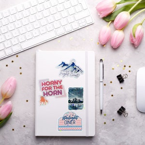 May include: A white notebook with colorful stickers on it. The stickers feature a mountain range with the text "Welcome to the Night Court", a pink and blue sticker with the text "Horny for the Horn", a blue and white sticker with the text "Welcome to the Night Court", and a pink and blue sticker with the text "Starlight Diner".