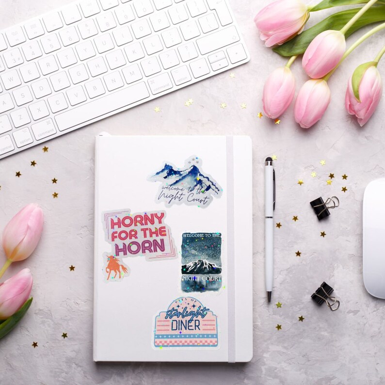May include: A white notebook with colorful stickers on it. The stickers feature a mountain range with the text "Welcome to the Night Court", a pink and blue sticker with the text "Horny for the Horn", a blue and white sticker with the text "Welcome to the Night Court", and a pink and blue sticker with the text "Starlight Diner".