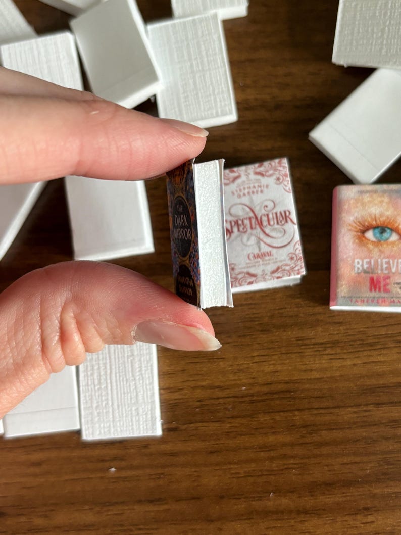 May include: A close-up of three miniature books with colorful covers. The books are titled "The Dark Mirror", "Spectacular", and "Believe Me".