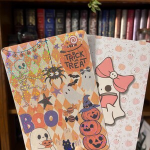 May include: Two Halloween-themed cards. One features a skeleton, spider, bats, ghosts, and the words "Trick or Treat" and "BOO!". The other card has a ghost with a pink bow and pumpkins. Both cards have a holographic finish.