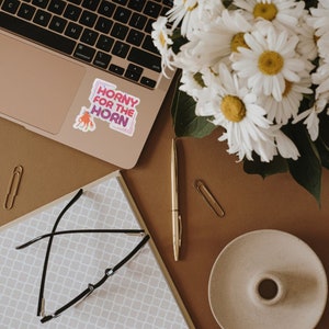 May include: A sticker with the text "Horny for the Horn" in pink and purple letters on a white background. The sticker is placed on a laptop computer with a brown background.