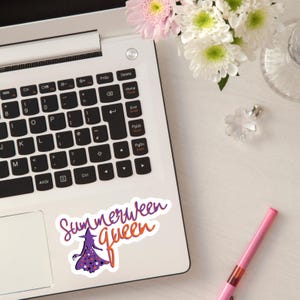 Może przedstawiać: Klawiatura laptopa z naklejką z napisem "Summerween Queen". Naklejka przedstawia fioletową wiedźmę z gwiazdami i tekst w kolorze pomarańczowym i fioletowym. Widoczny jest również różowy długopis i kwiaty.