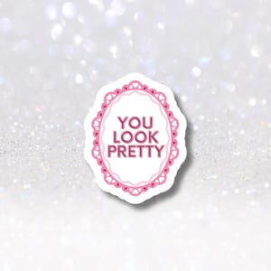Puede incluir: Una pegatina blanca con un marco ovalado rosa y las palabras "YOU LOOK PRETTY" en rosa. El marco tiene un borde festoneado. El fondo es blanco brillante.