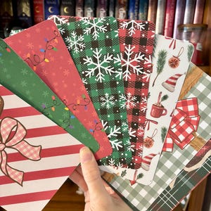 May include: A hand holds a fan of Christmas-themed cards. The cards feature various designs, including snowflakes, string lights, plaid patterns, and a bow. Colors include red, green, white, and brown. The cards are likely for gift tags or holiday greetings.