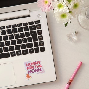 May include: A laptop computer with a sticker that says "Horny for the Horn" with a pink unicorn illustration. The sticker is on the right side of the laptop, near the edge.