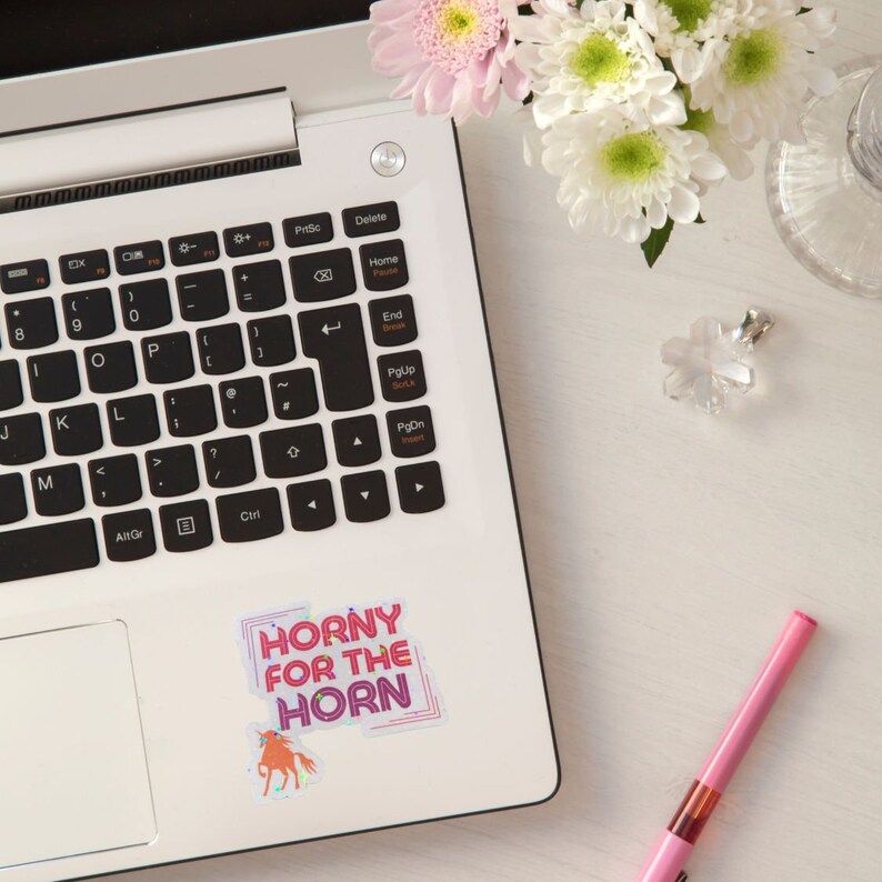 May include: A laptop computer with a sticker that says "Horny for the Horn" with a pink unicorn illustration. The sticker is on the right side of the laptop, near the edge.