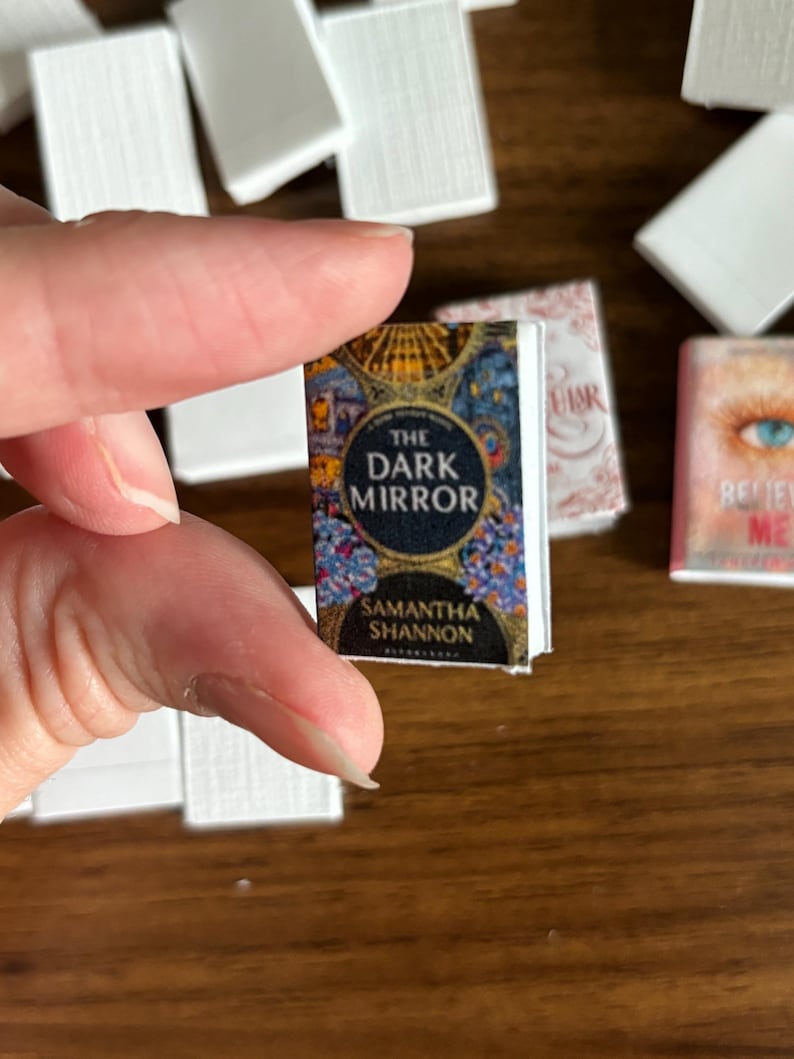 May include: A miniature paperback book with a colorful cover featuring a circular design and the title "The Dark Mirror" by Samantha Shannon.