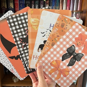 May include: A hand holds a fan of Halloween-themed cards. The cards feature orange, black, and white designs, including pumpkins, bats, bows, and a dog. Some cards have the letter "H" printed on them. The background includes a wooden bookshelf filled with books.