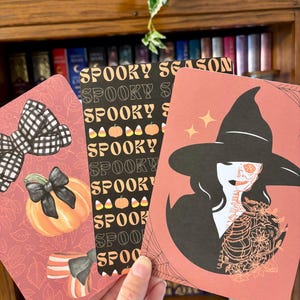 Könnte beinhalten: Drei Halloween-Karten. Eine Karte zeigt eine schwarz-weiße Schleife und einen Kürbis. Eine andere Karte trägt die Worte "Spooky Season" und Kürbisse. Die dritte Karte zeigt eine Hexe mit einem Skelettgesicht und einem Spinnennetz.