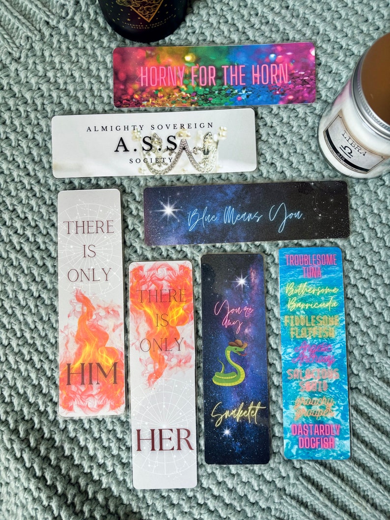 May include: A set of eight colorful bookmarks with various designs and text. The bookmarks feature a variety of themes, including a snake, a flame, and a crown. The text on the bookmarks includes phrases like "There is only him", "There is only her", "Blue means you", and "Horny for the horn".