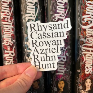 Puede incluir: Una pegatina blanca con los nombres Rhysand, Cassian, Rowan, Azriel, Ruhn y Hunt escritos en texto negro. La pegatina se sostiene frente a una fila de cuatro libros con el t&iacute;tulo "A Court of" en las espinas.