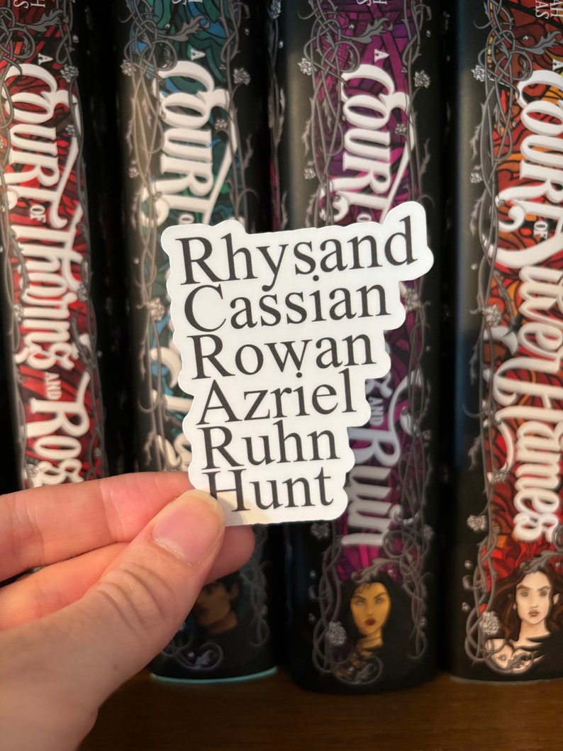 Puede incluir: Una pegatina blanca con los nombres Rhysand, Cassian, Rowan, Azriel, Ruhn y Hunt escritos en texto negro. La pegatina se sostiene frente a una fila de cuatro libros con el t&iacute;tulo "A Court of" en las espinas.