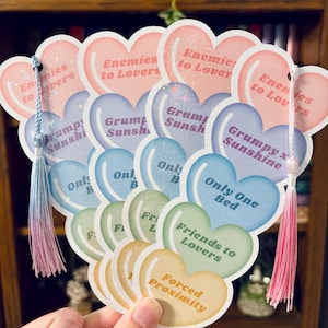 May include: A collection of heart-shaped bookmarks in various pastel colors, each with a different phrase like "Enemies to Lovers" and "Friends to Lovers." The bookmarks have tassels in shades of pink and blue.