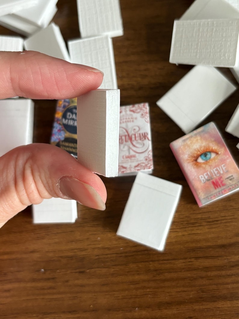 May include: A close-up of a stack of miniature white books. The books are all the same size and have a textured cover. The books are arranged in a stack, with the top book being held by a hand.
