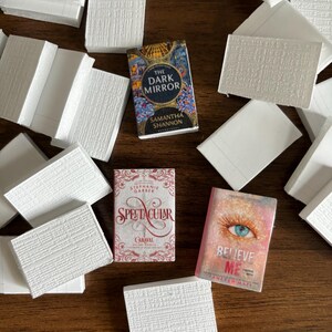 May include: A flat lay of several white books with three paperback books on top. The paperback books are titled "The Dark Mirror" by Samantha Shannon, "Spectacular" by Stephanie Garber, and "Believe Me" by Tahereh Mafi.