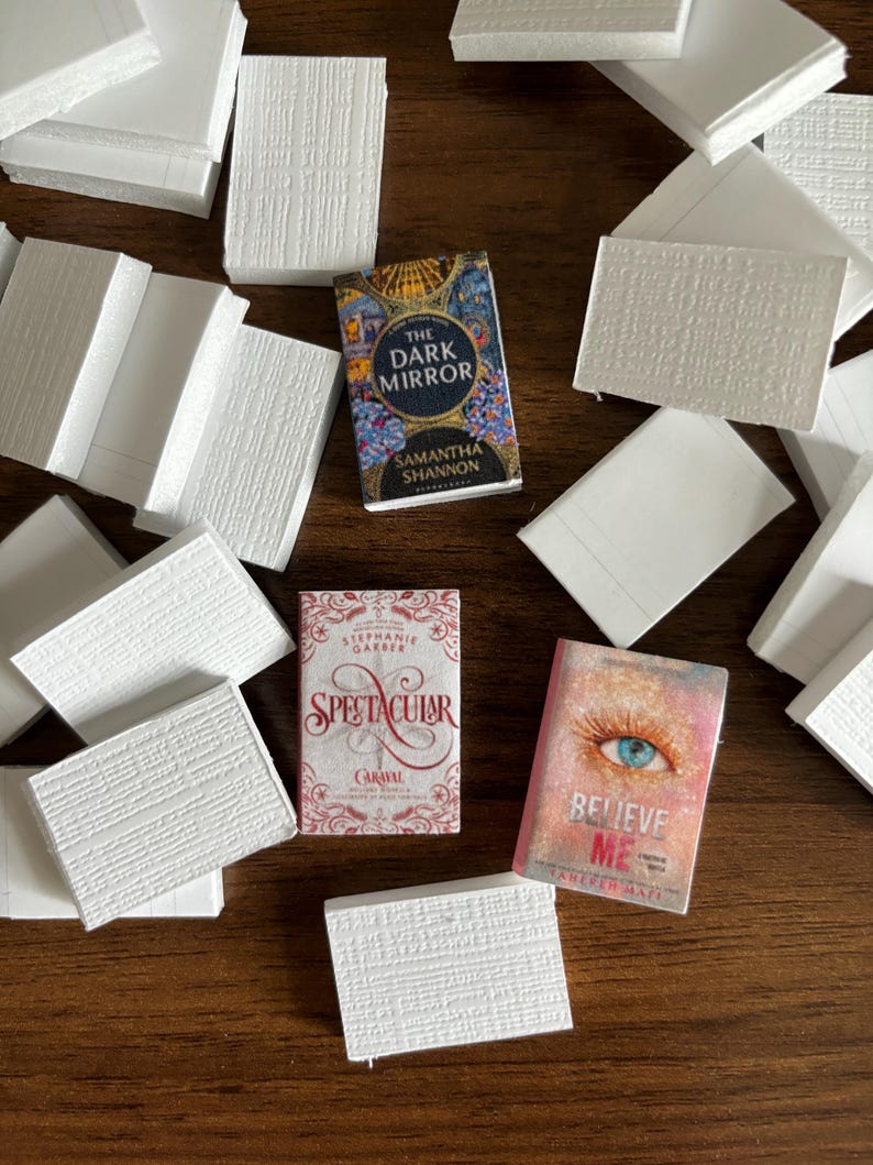 May include: A flat lay of several white books with three paperback books on top. The paperback books are titled "The Dark Mirror" by Samantha Shannon, "Spectacular" by Stephanie Garber, and "Believe Me" by Tahereh Mafi.