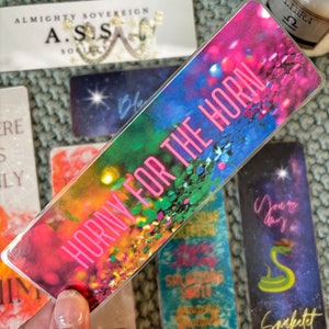 May include: A colorful bookmark with a rainbow background and the text "Horny for the Horn" in pink letters.