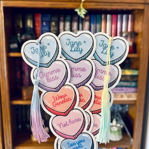 May include: A collection of heart-shaped bookmarks with pastel colors and text. The bookmarks have phrases like "Jane + Lily", "Gimme Kiss", "Wow Genetic", "Not Here", and "See you next season". Each bookmark has a tassel.