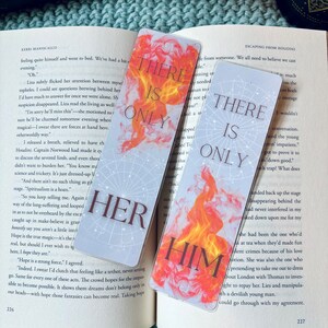 May include: Two bookmarks with a fiery orange and yellow design. The text on the bookmarks reads "There is only" and "There is only" with the words "her" and "him" in large, bold letters below the text. The bookmarks are on a blue knitted surface.