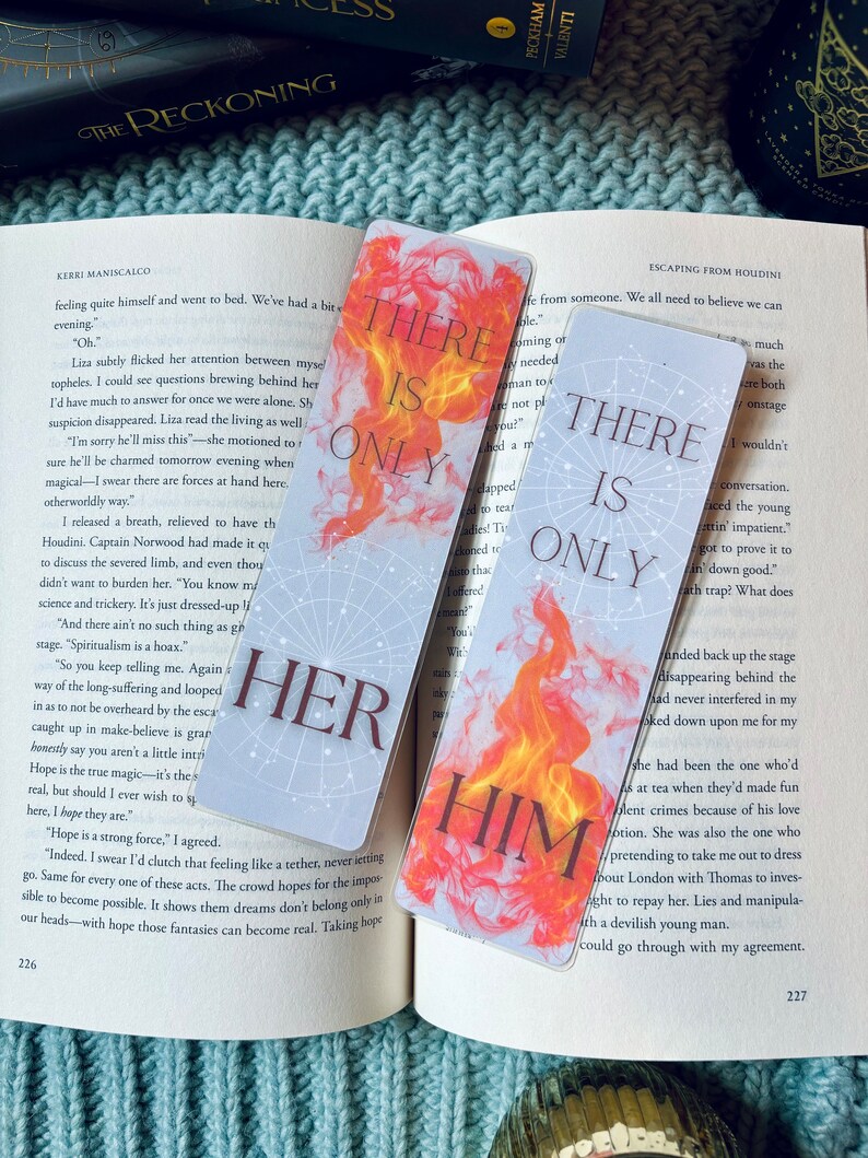 May include: Two bookmarks with a fiery orange and yellow design. The text on the bookmarks reads "There is only" and "There is only" with the words "her" and "him" in large, bold letters below the text. The bookmarks are on a blue knitted surface.